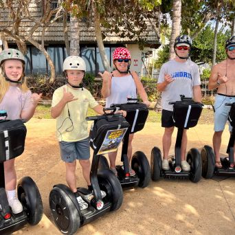 Group of five people on Segways, wearing helmets, smiling and gesturing, with trees and a building in the background.