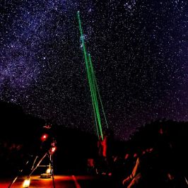 Night sky with stars and green laser pointing up from observatory with people and equipment.