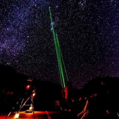 Night sky with stars and green laser pointing up from observatory with people and equipment.