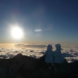 Two people in jackets sitting on a mountaintop above clouds at sunrise.