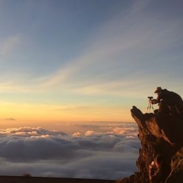 Person photographing sunrise above clouds from rocky cliff.