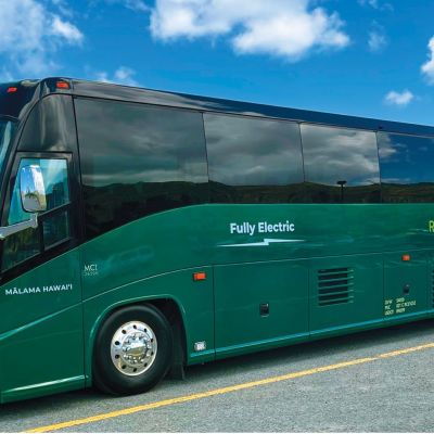 Green fully electric tour bus parked on road with clear blue sky backdrop.
