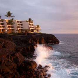 Sunset view of a seaside hotel with palm trees on rocky cliffs by the ocean.