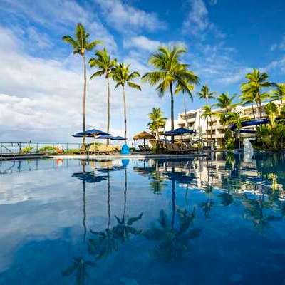 Poolside view with palm trees and hotel under a blue sky with scattered clouds.
