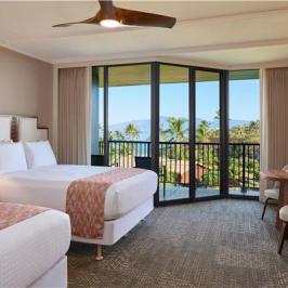 Hotel room with two beds, balcony view, ceiling fan, and desk.