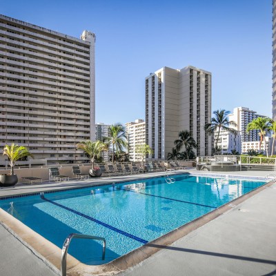 Rooftop pool area with lounge chairs, surrounded by tall buildings and palm trees under a clear blue sky.