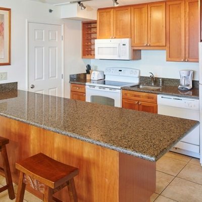 Modern kitchen with granite island, two stools, wooden cabinets, and white appliances.