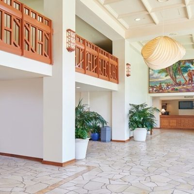 Hotel lobby with stone floor, wooden balcony, potted plants, and a reception desk.