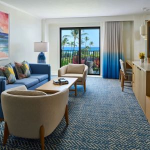 Modern living room with blue sofa, armchairs, painting, and ocean view through large window.