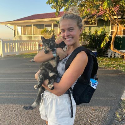 Person smiling, holding a cat on a sunny day near a house.