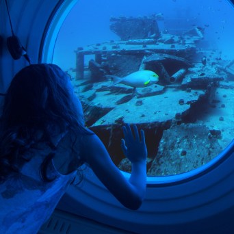 Child looks out submarine window at sunken ship and fish.