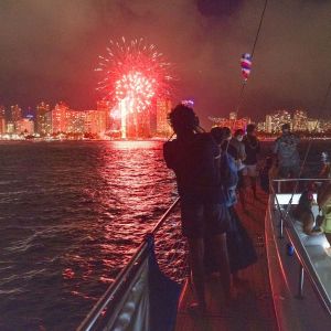 People on boat watching fireworks over city skyline at night.