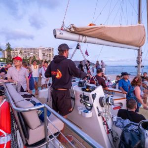 People gathered on a sailboat at sunset, with buildings and palm trees in the background.