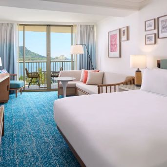 Hotel room with a bed, sofa, and balcony view of a mountain and ocean.