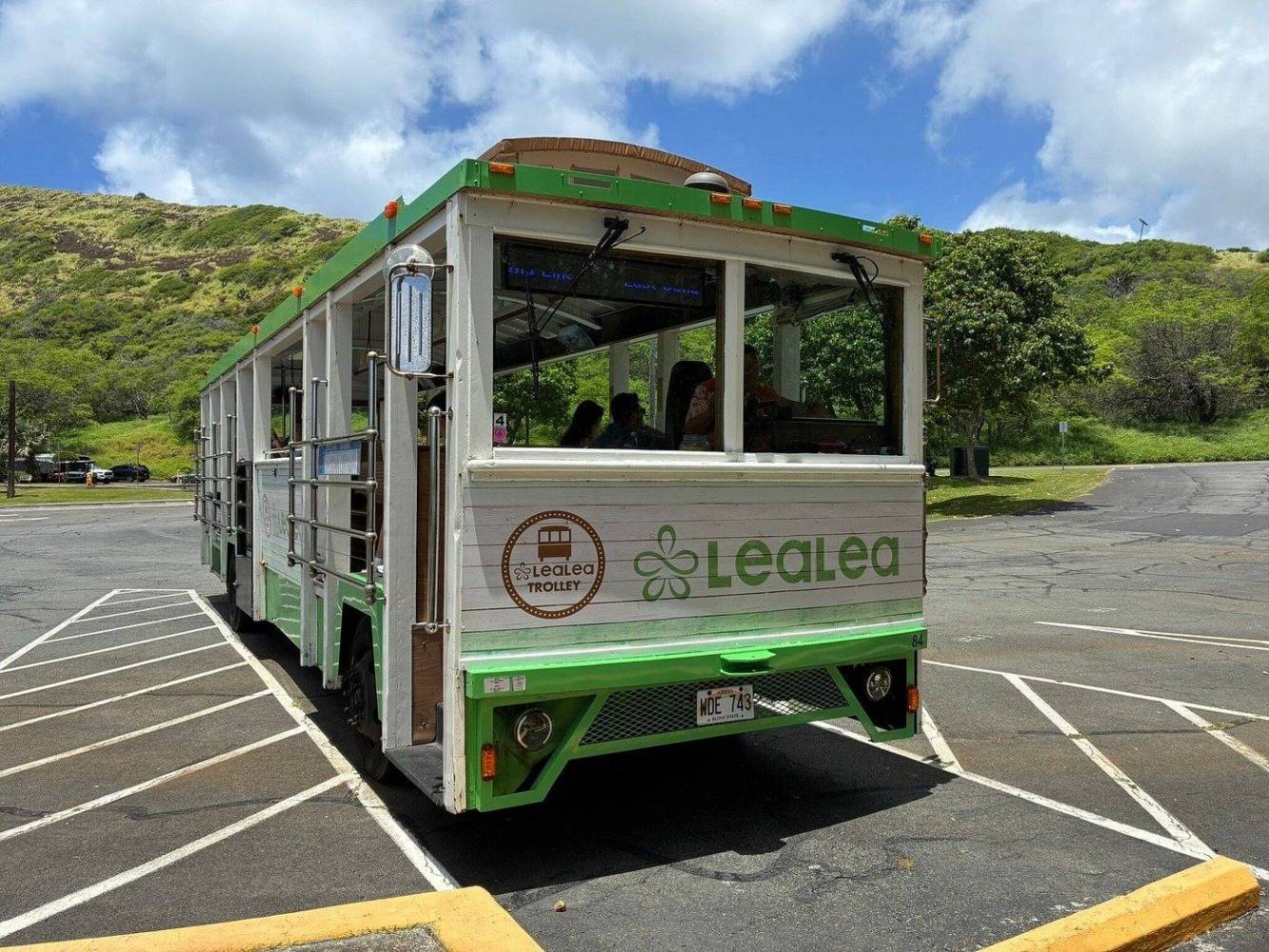 Green and white open-air trolley parked in a lot with hills in the background.