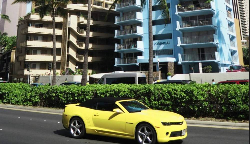 Yellow convertible car driving past palm trees and a blue building.