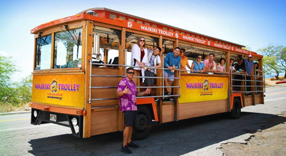 People standing and sitting on an orange Waikiki trolley on a sunny day.