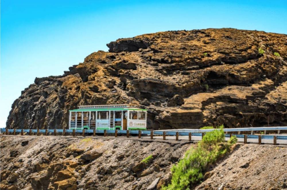 Tram on a road beside a rocky hill under a clear blue sky.