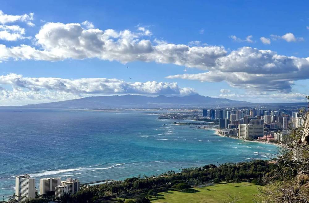 Coastal cityscape with ocean, high-rise buildings, and distant mountains under a partly cloudy sky.