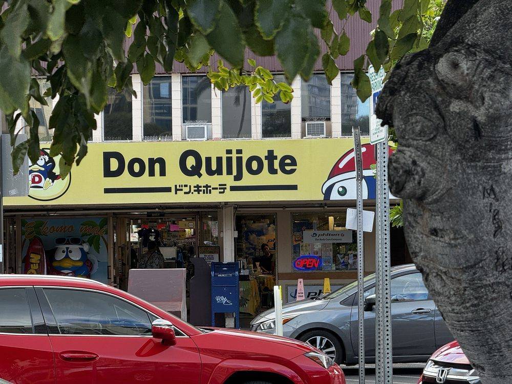 Storefront with 'Don Quijote' sign above, tree and parked cars in foreground.