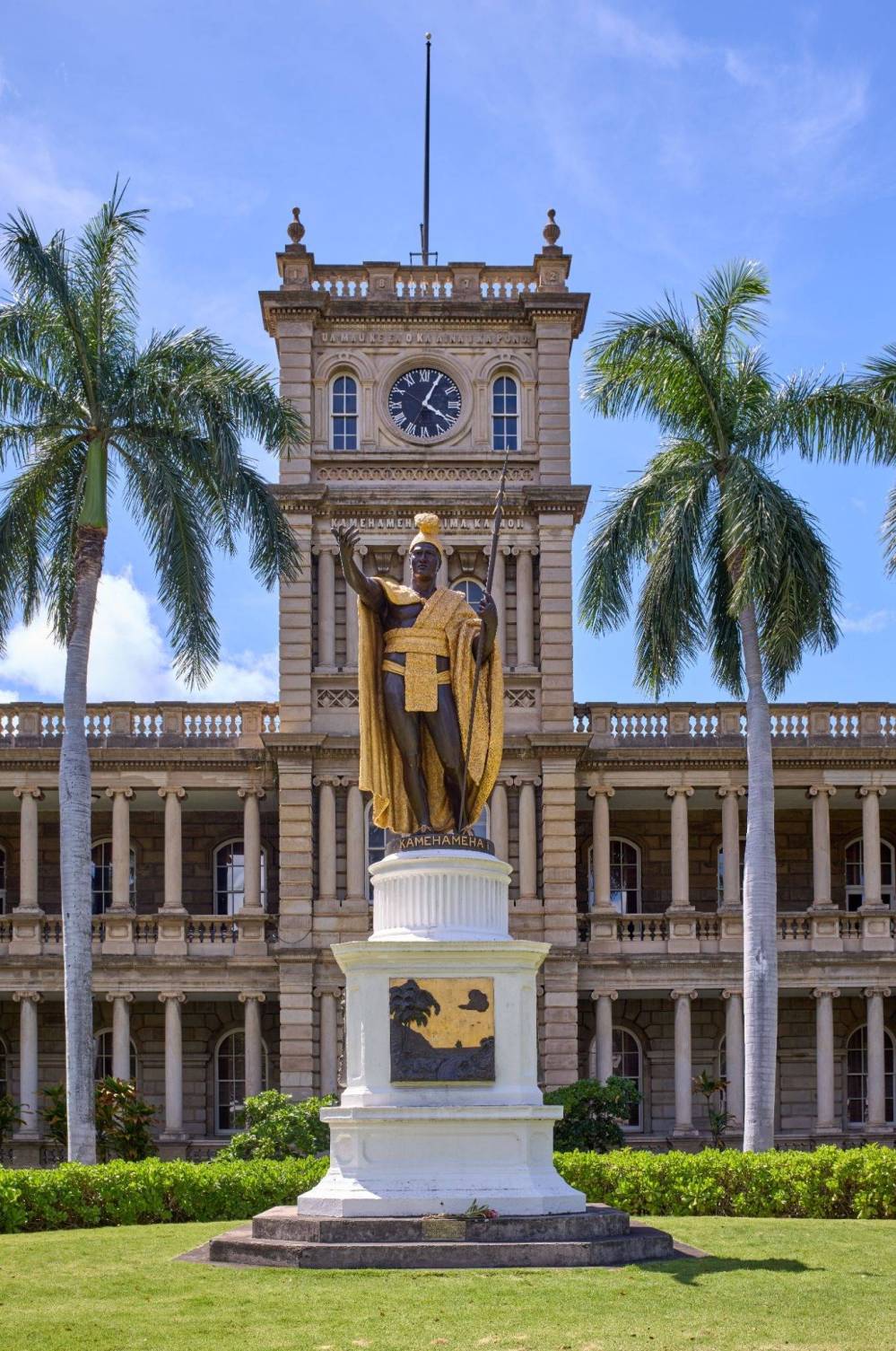 Statue of a robed figure in front of a historic building with a clock, flanked by palm trees.