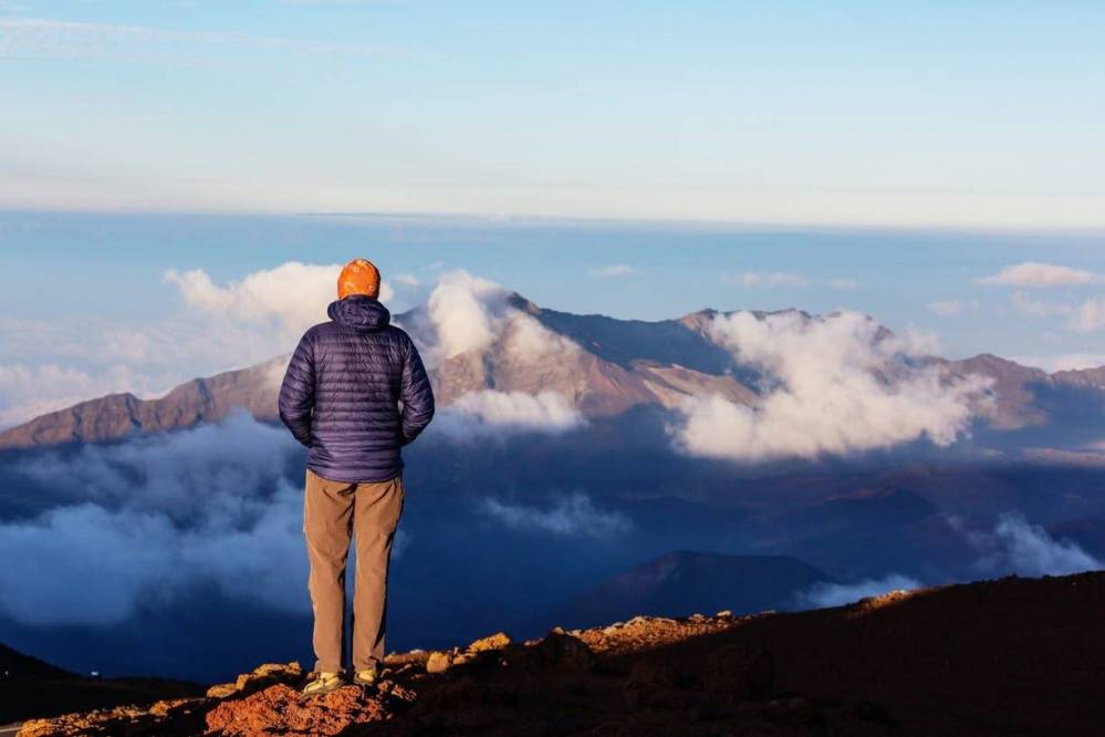 Person in jacket and hat stands on rocky hill, overlooking clouds and distant mountain.