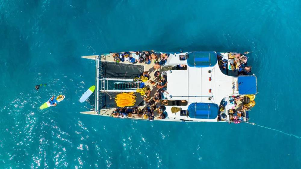 Aerial view of a boat with people on a blue ocean; a kayak and swimmer nearby.