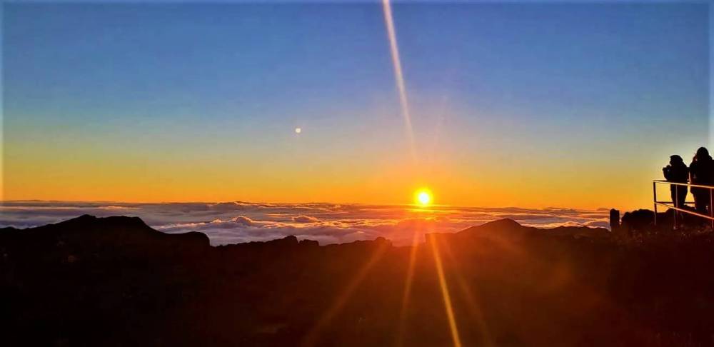 Silhouetted people watch a sunrise over a mountain landscape.