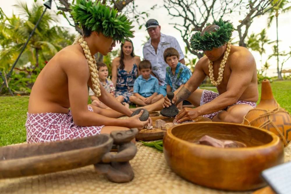 Two people in traditional attire preparing food outdoors, with a family watching.