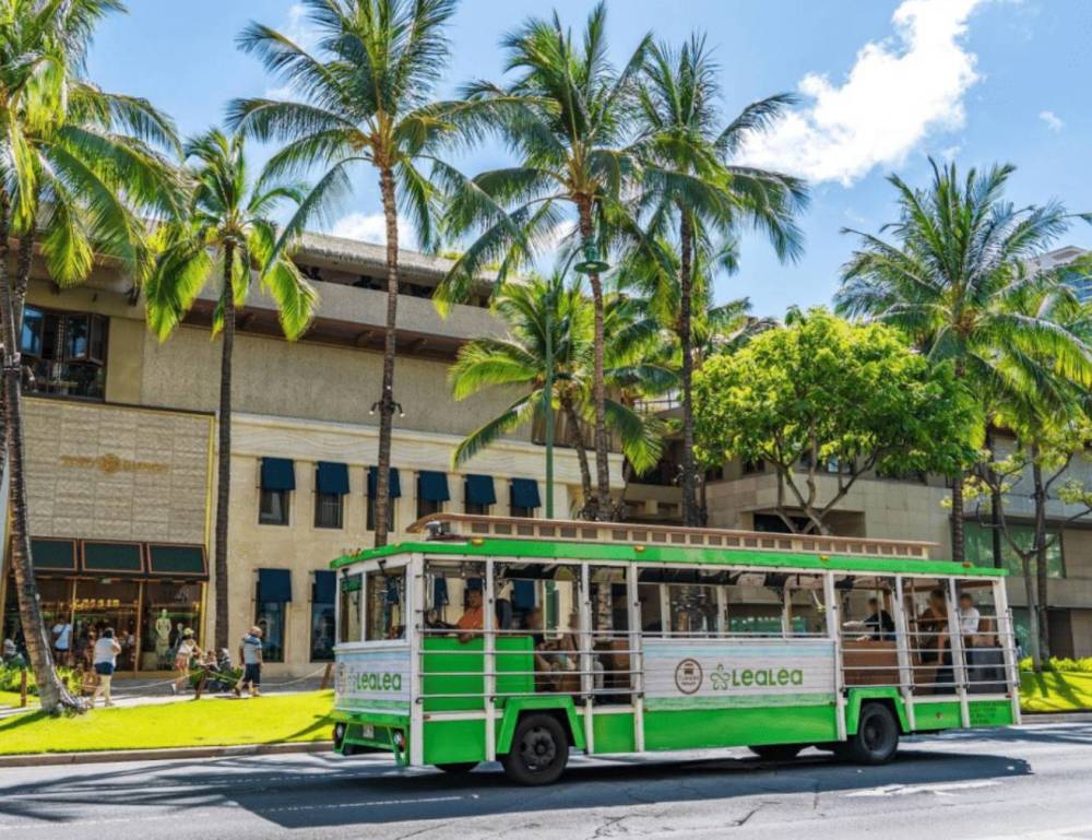 Green trolley on street with palm trees and building in background.