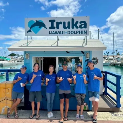 Six people in blue shirts pose in front of a dolphin tour kiosk at a marina.